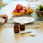 Recovery Nutrition - From above of crop anonymous person sitting at table with pile of various medicines and bowls of fresh lettuce salad and ripe fruits in kitchen