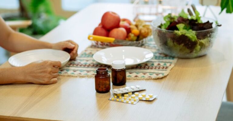 Recovery Nutrition - From above of crop anonymous person sitting at table with pile of various medicines and bowls of fresh lettuce salad and ripe fruits in kitchen