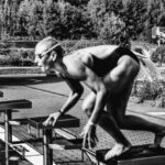 Swim Starts - Black and white side view full body sporty swimmer in swimming suit and goggles standing on block in track start position preparing to dive in outside pool