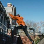 Brick Workouts - Side view full body fit male tracer wearing orange hoodie jumping from brick parapet towards building railing on clear day