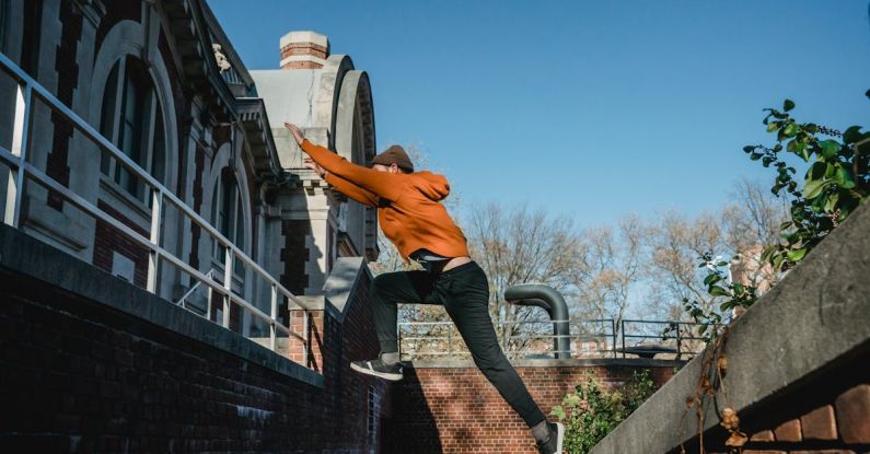 Brick Workouts - Side view full body fit male tracer wearing orange hoodie jumping from brick parapet towards building railing on clear day